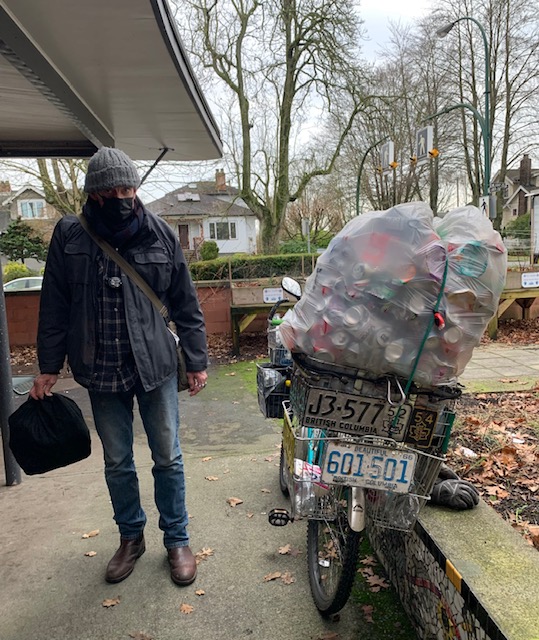 A man in jeans, a dark jacket, toque and wearing a mask stands next to his bicycle which has baskets full of can on it. Each basket is adorned with license plates in different styles.