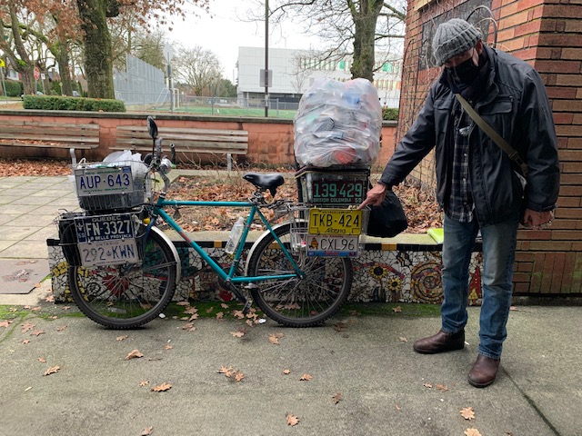 A man in a dark jacket, toque and jeans points at the license plates that are displayed on the front and rear baskets of his blue bicycle.