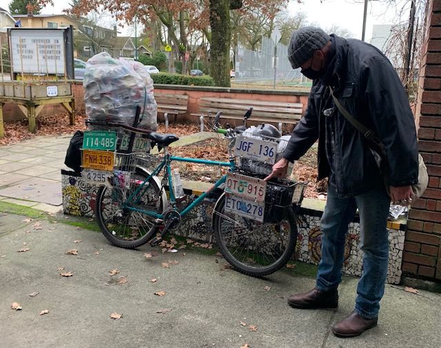 A man in a dark jacket, toque and jeans points at the license plates that are displayed on the front and rear baskets of his blue bicycle.